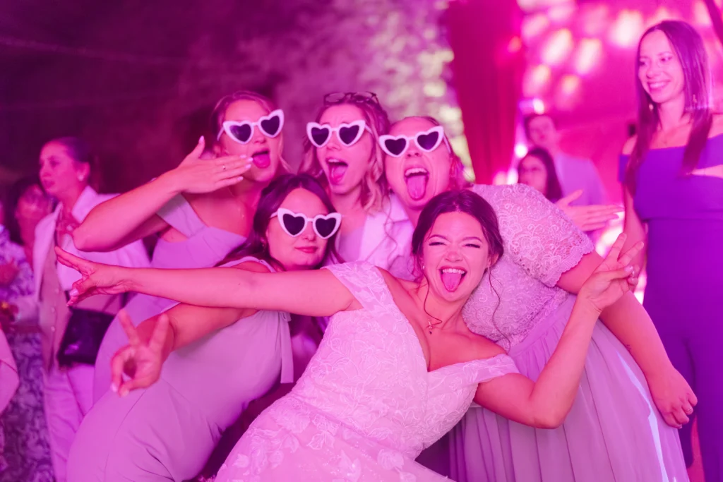 Mariée et ses amies posant avec des lunettes en forme de cœur en soirée, photographie de mariage fun et décalée par Studio Timeless.