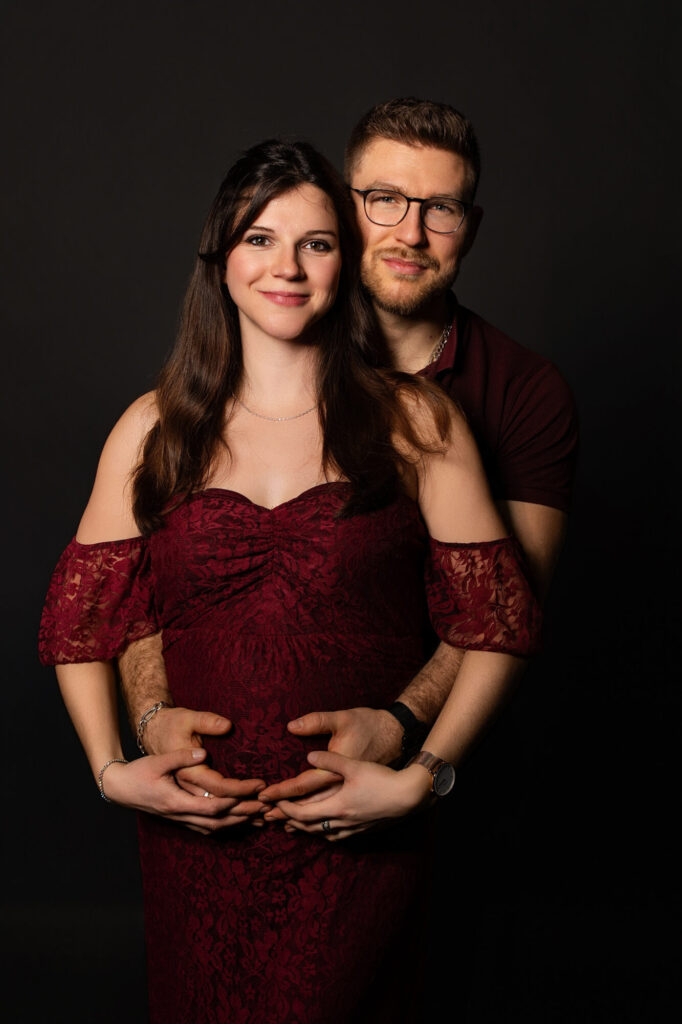 Couple en séance maternité, futur père enlace par derrière sa compagne enceinte, leurs mains formant un cœur sur le ventre rond, tous deux souriants, robe dentelle bordeaux – Limoges, Studio Timeless