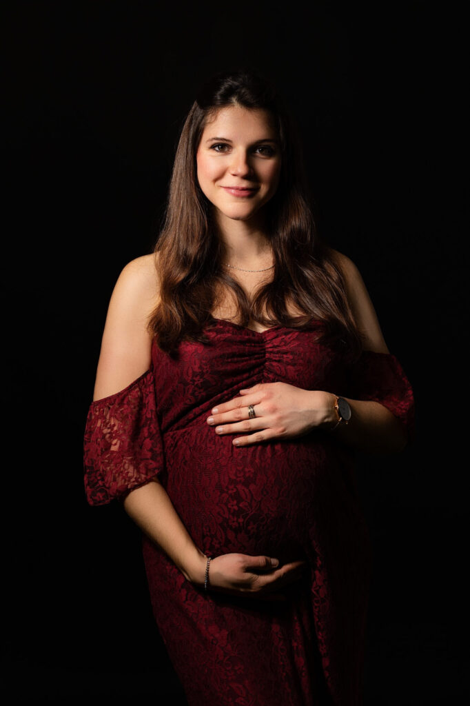 Femme enceinte en robe dentelle bordeaux souriante, mains posées sur son ventre arrondi, bague visible, regard chaleureux et naturel lors d'une séance maternité à Limoges – Studio Timeless