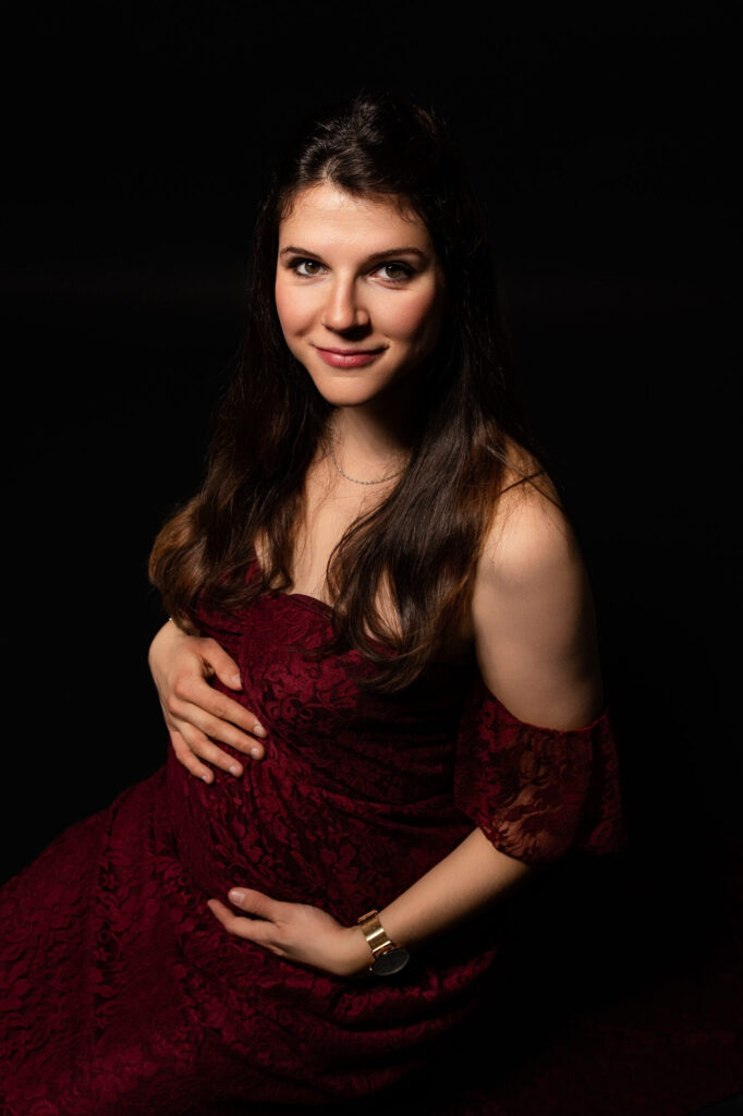 Femme enceinte élégante en robe velours bordeaux foncé, mains posées sur son ventre arrondi, regard direct et confiant face à la caméra, portrait glamour – séance maternité à Limoges, Studio Timeless