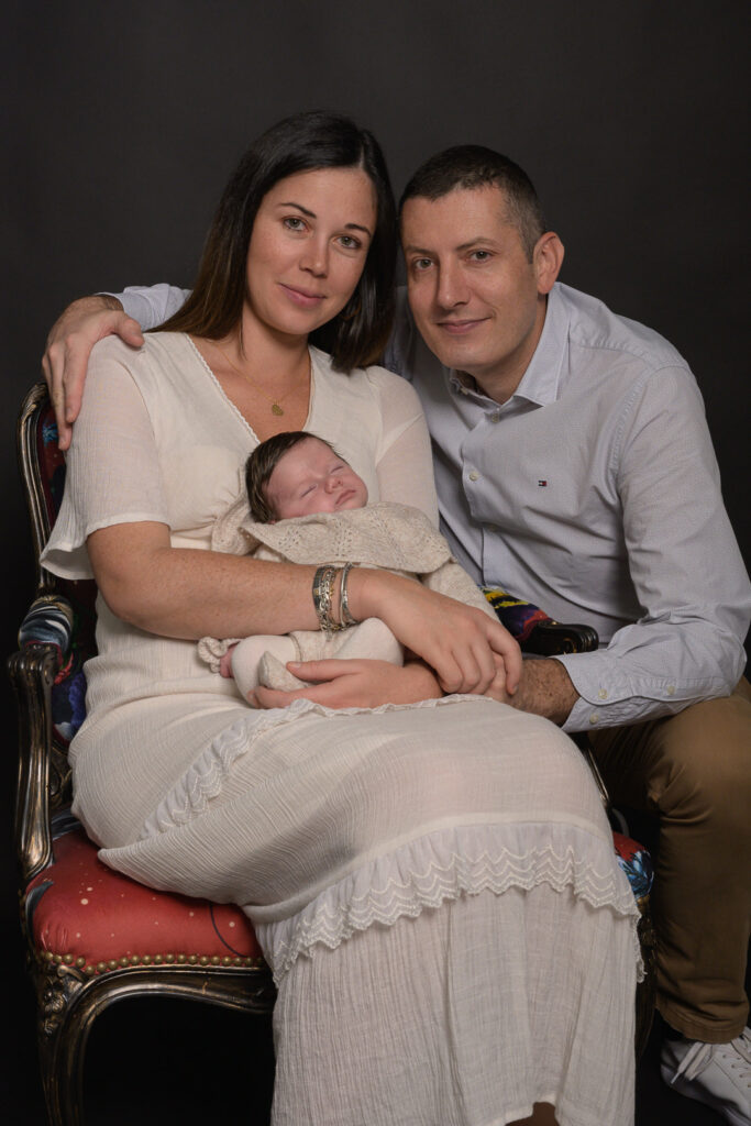 Portrait de famille avec maman en robe blanche et papa souriant tenant leur nouveau-né dans un fauteuil fleuri lors d'une séance photo naissance à Limoges – Studio Timeless