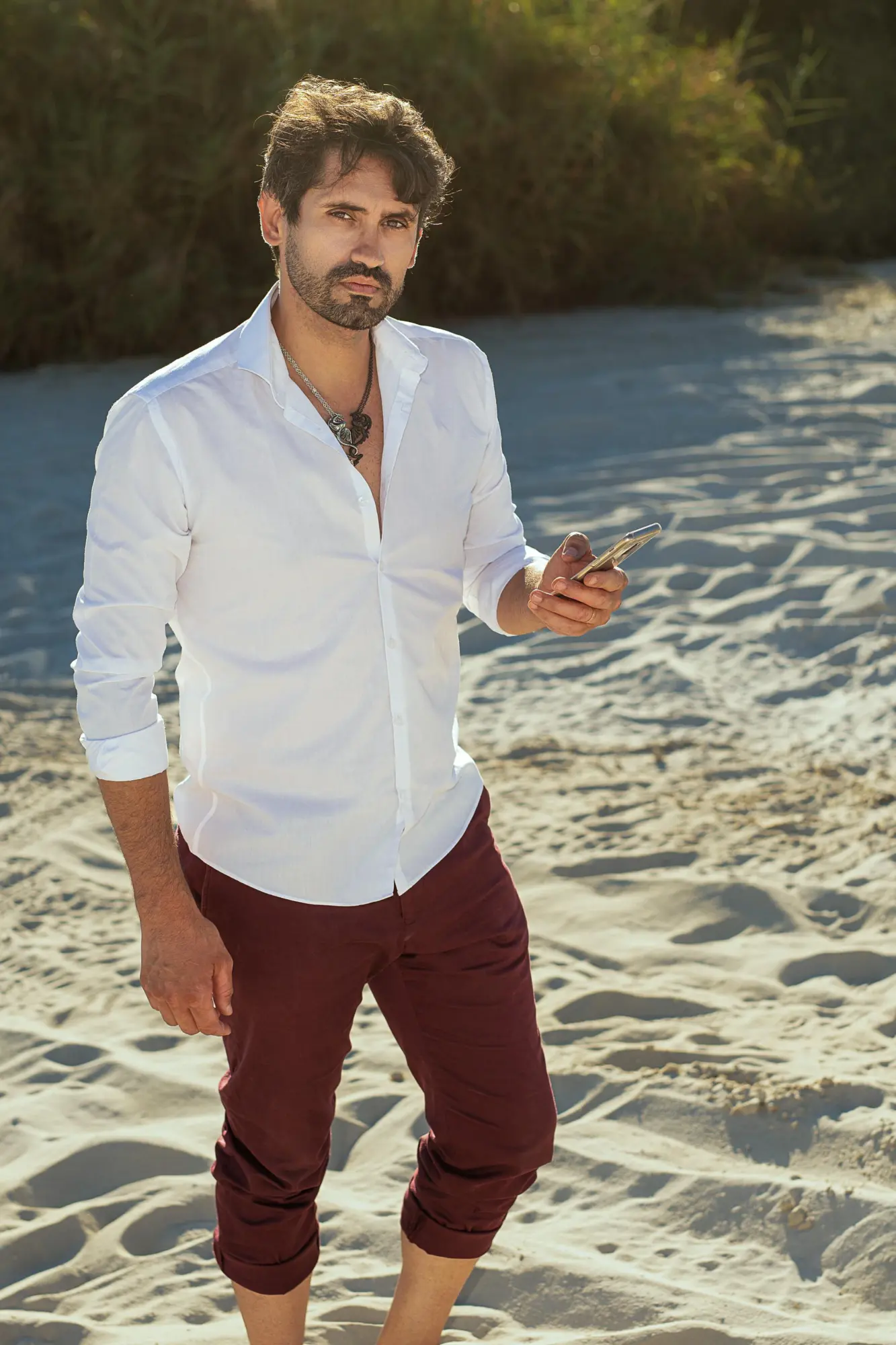 Portrait masculin en extérieur d'un homme élégant en chemise blanche et pantalon bordeaux, pieds dans le sable avec téléphone, pose décontractée – séance photo portrait à Limoges, Studio Timeless